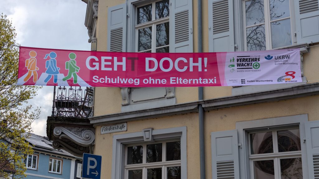 An einem Gebäude hängt ein Transparent mit der Aufschrift "GEHT DOCH! Schulweg ohne Elterntaxi" mit Ikonen von gehenden Kindern, die die Schüler auffordern, zu Fuß zur Schule zu gehen, anstatt mit dem Auto zu fahren. Auf der rechten Seite befinden sich Logos von Sponsoren.