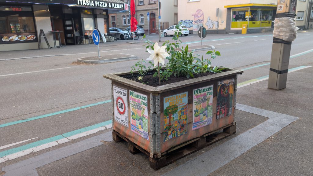 Ein hölzerner Blumenkasten mit grünen Pflanzen und einer großen weißen Blume steht an einer Straßenecke. Der Kasten ist mit bunten Plakaten beklebt, und im Hintergrund sind Geschäfte und Gebäude zu sehen.