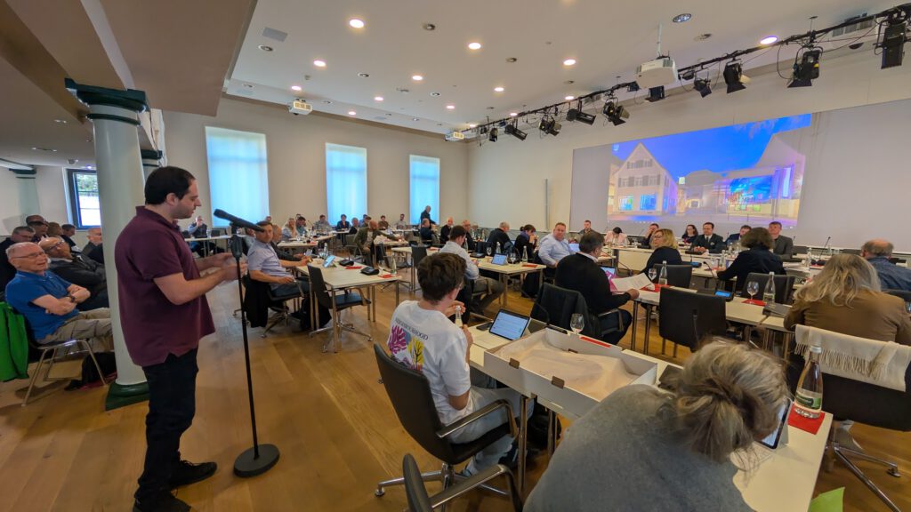 Ein Mann spricht an einem Mikrofon in einem großen, modernen Konferenzraum, in dem viele Menschen an Tischen sitzen. An der Wand hinter den Zuhörern ist ein großes projiziertes Bild eines Gebäudes zu sehen.