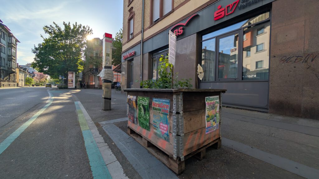 Ein hölzerner Pflanzkasten mit Grünpflanzen steht an einem sonnigen Tag in einem städtischen Gebiet auf einem Gehweg in der Nähe eines Fahrradwegs, neben einem Gebäude mit großen Fenstern und bunten Plakaten.