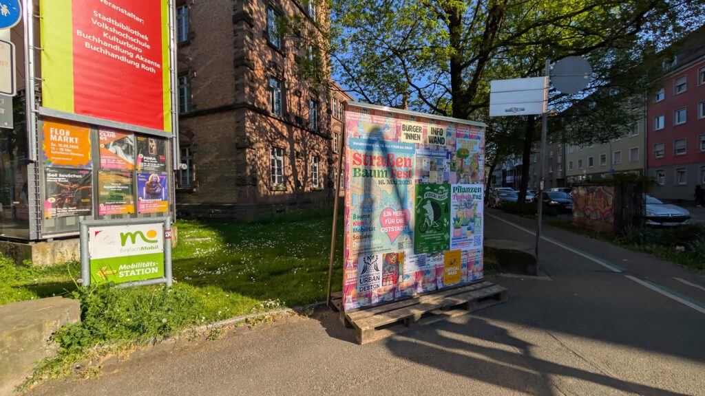 Ein Bürgersteig in einer Stadt mit einer großen Tafel, die mit bunten Plakaten bedeckt ist, eine Grasfläche mit Schildern auf der linken Seite und Gebäude im Hintergrund unter belaubten Bäumen an einem sonnigen Tag.