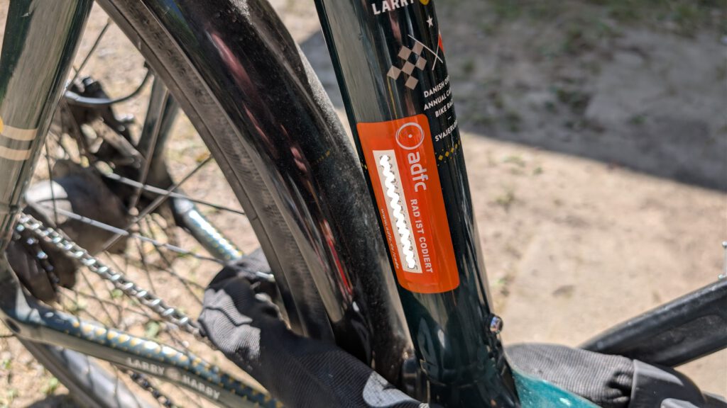 Nahaufnahme eines Fahrradrahmens mit einem orangefarbenen adfc Rad ist korrekt-Aufkleber und einem Kettenschloss. Der Boden darunter ist sonnenbeschienen, im Hintergrund sind Schatten und Gras zu sehen.