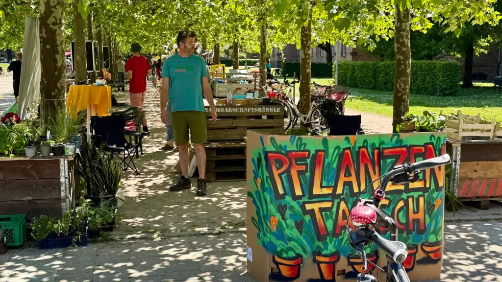 Ein Mann steht an einem von Bäumen beschatteten Marktstand im Freien, an dem verschiedene Pflanzen und Gegenstände angeboten werden. Davor steht ein buntes Schild mit der Aufschrift Pflanzentausch. Im Vordergrund ist ein Fahrrad geparkt.