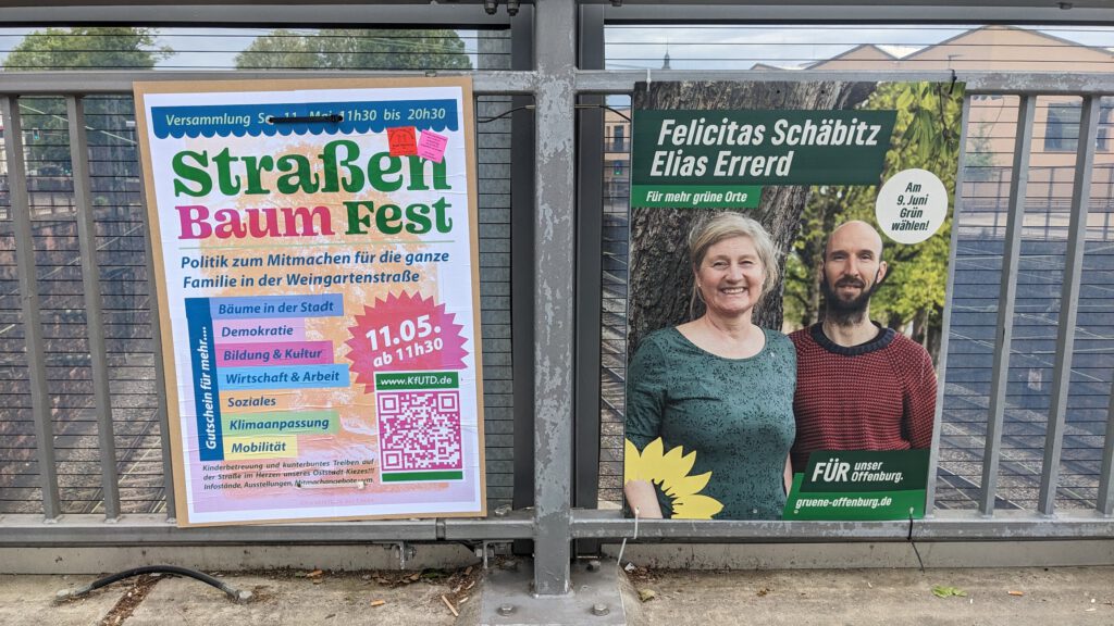 Zwei Plakate aus 2024 an einem Metallzaun: Das eine wirbt mit buntem Text und einem QR-Code für ein Straßenbaumfest, das andere zeigt zwei Personen, Felicitas Schäbitz und Elias Errerd, die mit grünem Hintergrund für die Grüne Partei werben.
