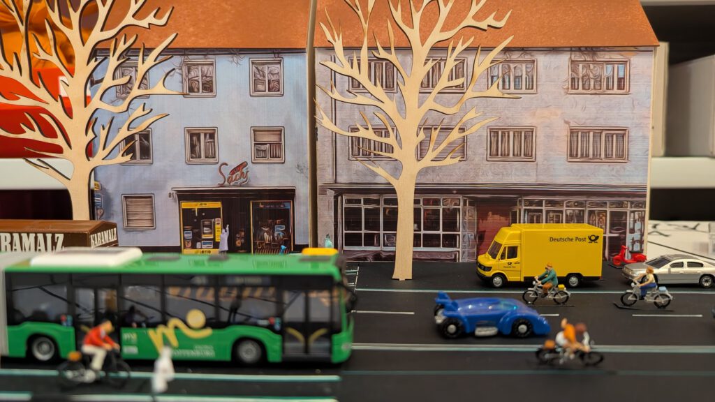 Eine Miniaturstadtszene zeigt Modellfahrzeuge, darunter einen grünen Bus, einen gelben Lieferwagen, ein blaues Auto und mehrere Motorräder, vor Papierbäumen und einer Kulisse aus illustrierten Gebäudefassaden.