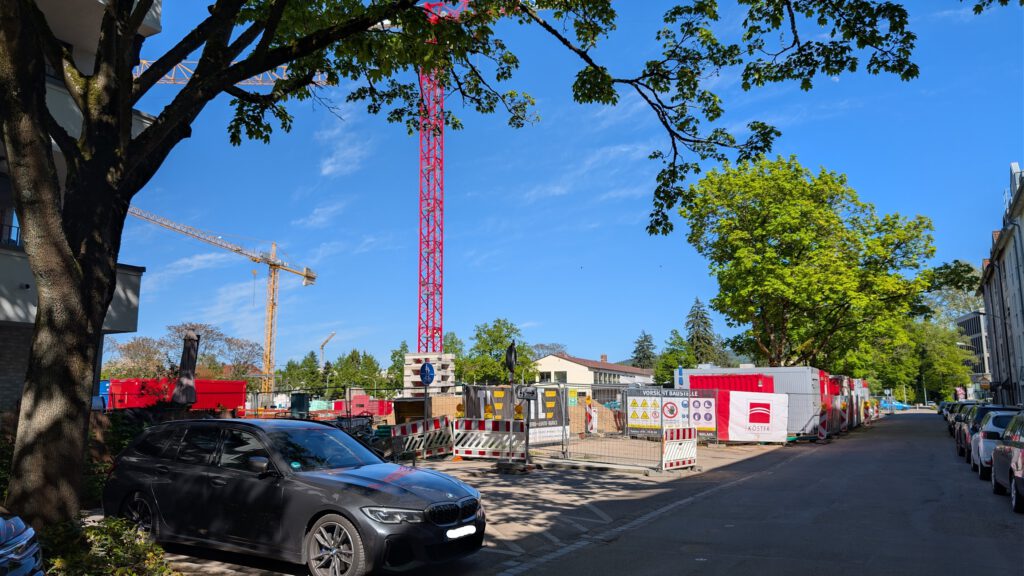 Eine Straße neben einer Baustelle mit Kränen, rot-weißen Absperrungen und tragbaren Gebäuden unter einem strahlend blauen Himmel. Geparkte Autos säumen die Straße, und Bäume spenden Schatten.