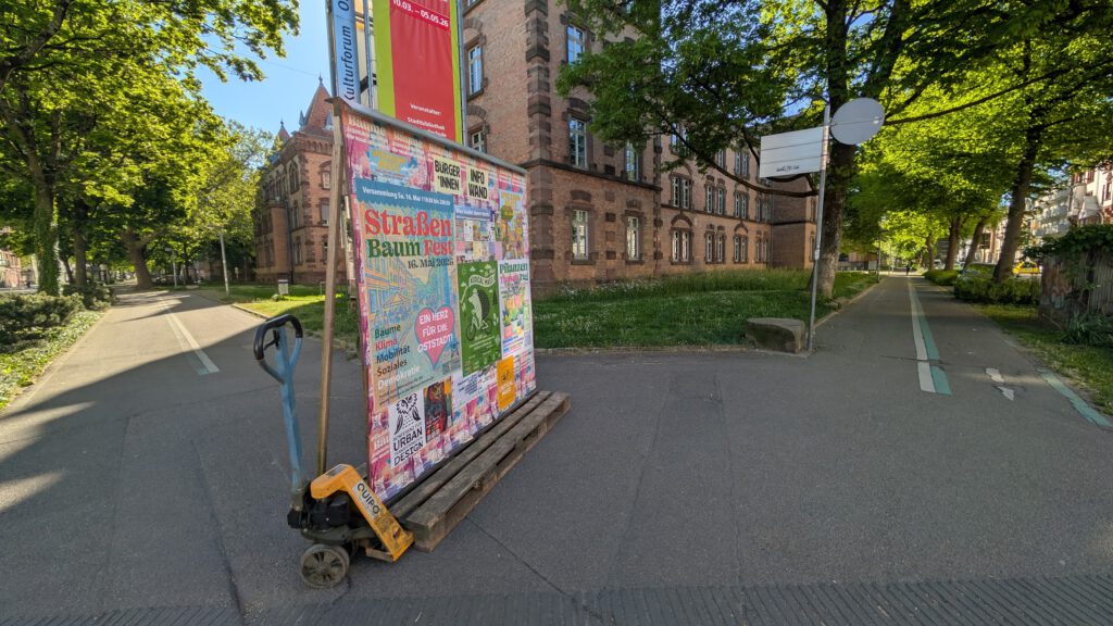 Colorful poster board on a pallet cart in a tree-lined urban street with a red brick building in the background
