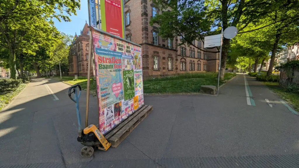 Colorful poster board on a pallet cart in a tree-lined urban street with a red brick building in the background