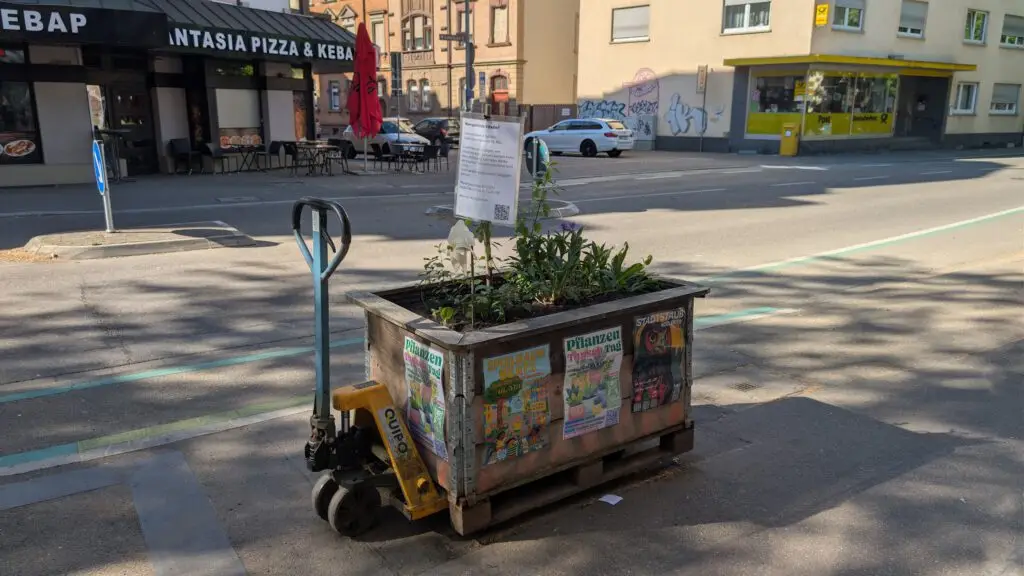 Ein großer Blumenkübel mit verschiedenen Pflanzen steht auf einem Hubwagen am Rande einer städtischen Straße. Plakate und Aufkleber bedecken die Pflanzschale, und im Hintergrund sind Restaurants und geparkte Autos zu sehen.