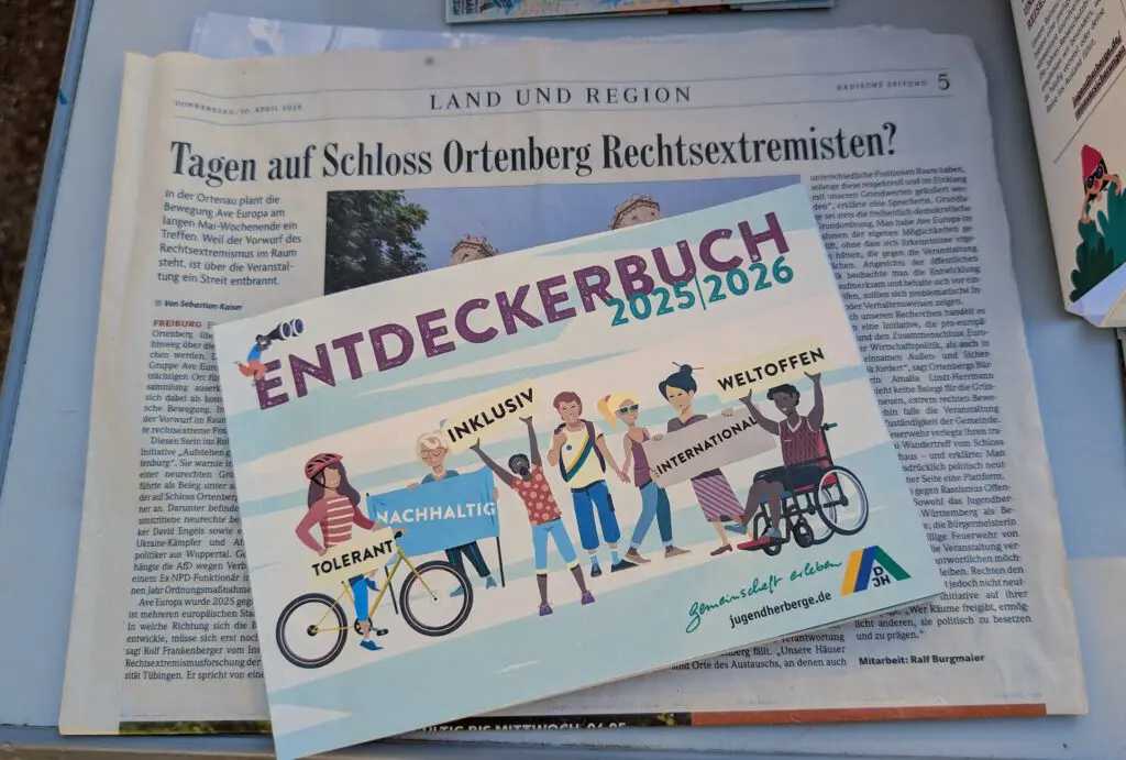 Eine farbenfrohe Broschüre mit dem Titel Entdeckerbuch 2025/2026 mit Abbildungen verschiedener Kinder liegt auf einer deutschen Zeitung mit der Schlagzeile Tage auf Schloss Ortenberg Rechtsextremisten?.