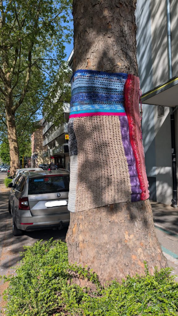 Ein großer Baumstamm auf einer Straße in der Stadt ist in einen bunten gestrickten oder gehäkelten Stoff mit Streifen in Blau, Rot, Lila, Beige und Grau gehüllt. Im Hintergrund sind geparkte Autos und Gebäude zu sehen.
