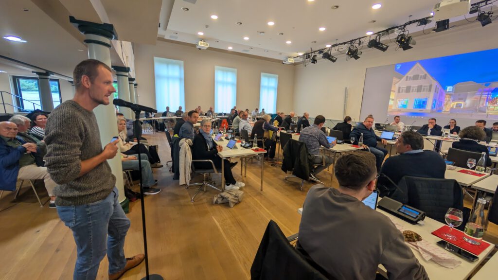 Michael Scherwitz spricht in ein Mikrofon im Salmen, in dem Stadtverwaltung und Gemeinderat an Tischen sitzen, einige mit Laptops. Auf einem großen Bildschirm wird ein Bild vom Salmen im vorderen Teil des Raums angezeigt.