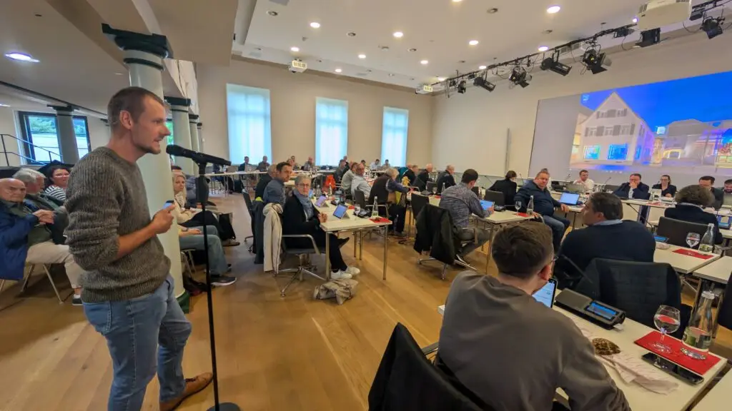Michael Scherwitz spricht in ein Mikrofon im Salmen, in dem Stadtverwaltung und Gemeinderat an Tischen sitzen, einige mit Laptops. Auf einem großen Bildschirm wird ein Bild vom Salmen im vorderen Teil des Raums angezeigt.