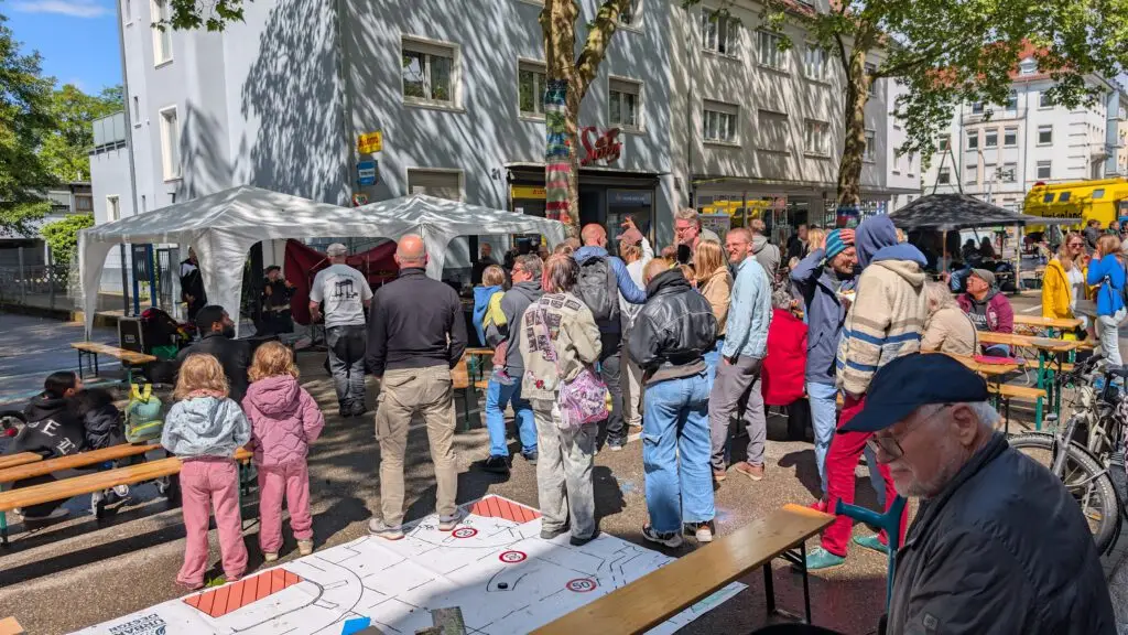 Eine Gruppe von Menschen versammelt sich im Freien in einer sonnigen städtischen Umgebung mit Bänken, Zelten und Spielen. Kinder und Erwachsene interagieren, einige stehend und einige sitzend, in der Nähe von Gebäuden und Bäumen auf einer belebten Straße.