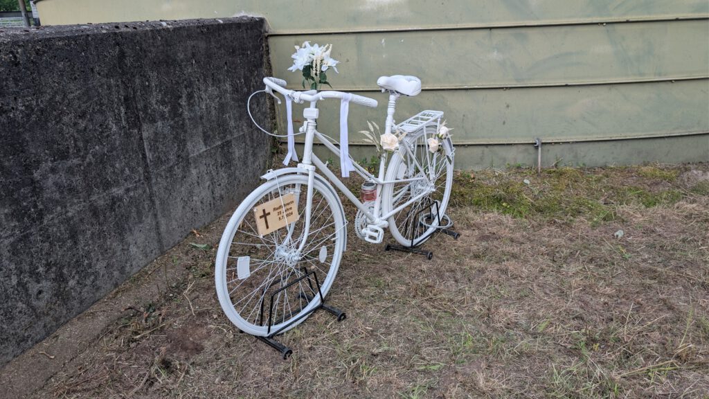 White bicycle painted white, decorated with white flowers and ribbons, leaning against a green wall; a wooden memorial plaque with a cross hangs from the front wheel.