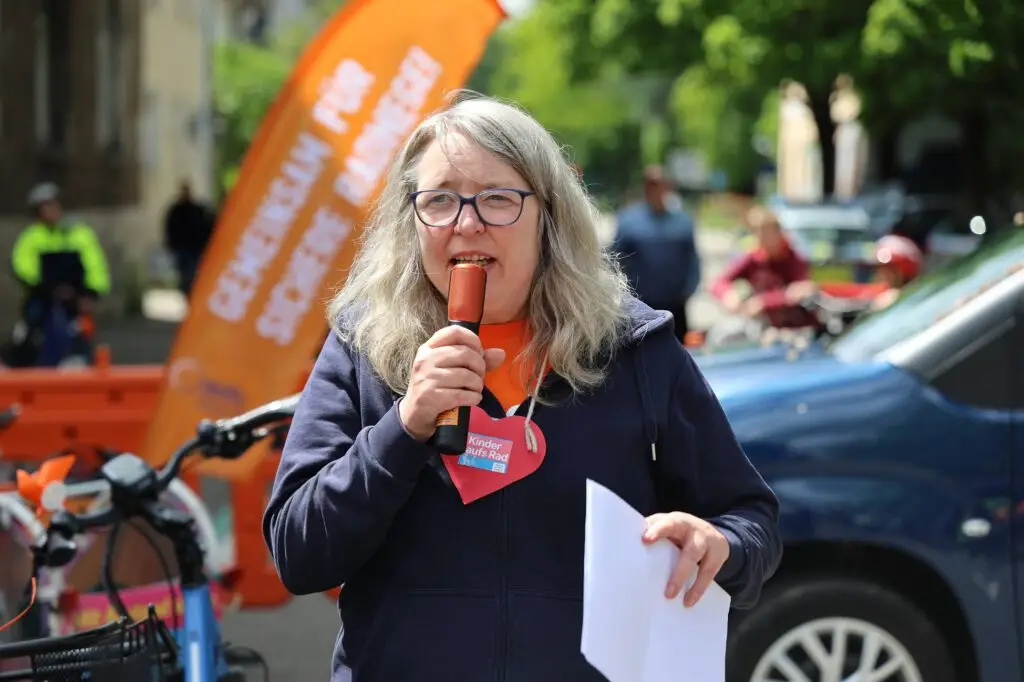 Eine Frau mit grauem Haar und Brille spricht bei einer Veranstaltung im Freien in ein Mikrofon. Sie hält Papiere in der Hand und steht vor orangefarbenen Bannern und einem geparkten Auto. Im Hintergrund sind Menschen und Fahrräder zu sehen.