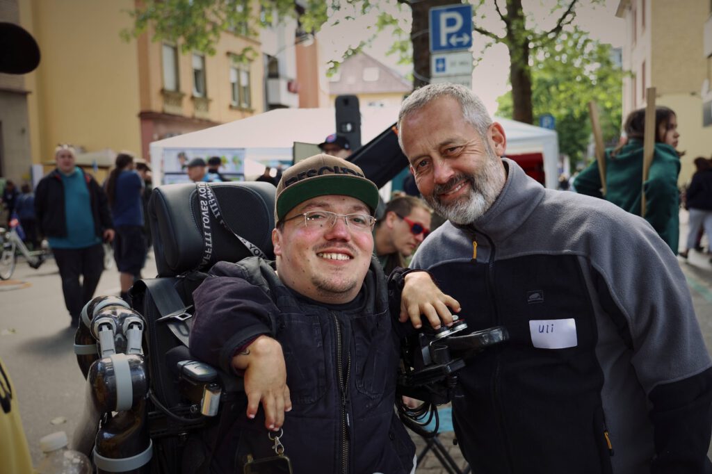 Ein Mann im Rollstuhl lächelt breit, sein Arm stützt sich auf einen Mann mit grauem Bart und Fleecejacke. Sie stehen draußen.