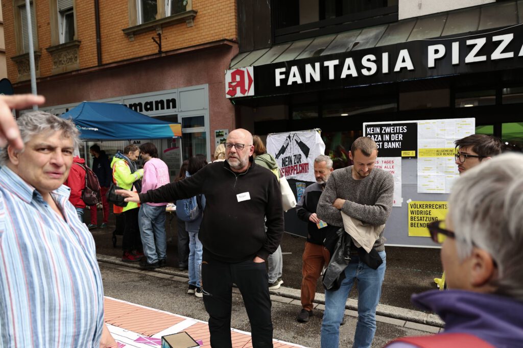 Menschen versammeln sich vor einer Pizzeria, einige halten Schilder in der Hand. Ein Mann in einem schwarzen Sweatshirt trägt ein Namensschild mit der Aufschrift Ralph.