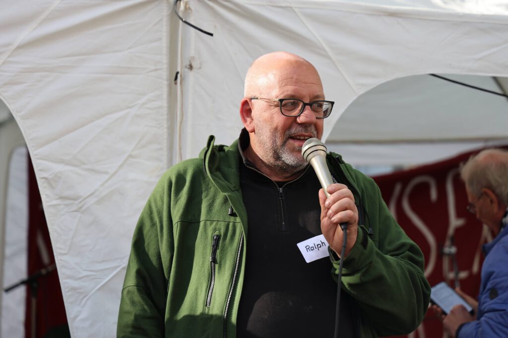 Glatzköpfiger Mann mit Brille, der eine grüne Jacke trägt und in ein Mikrofon spricht. Auf seinem Hemd befindet sich ein Namensschild mit der Aufschrift Ralph.