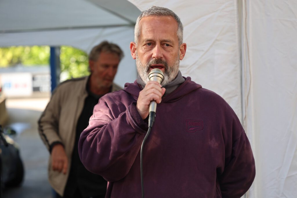 Man with gray beard and maroon hoodie speaks into a microphone under a white canopy.