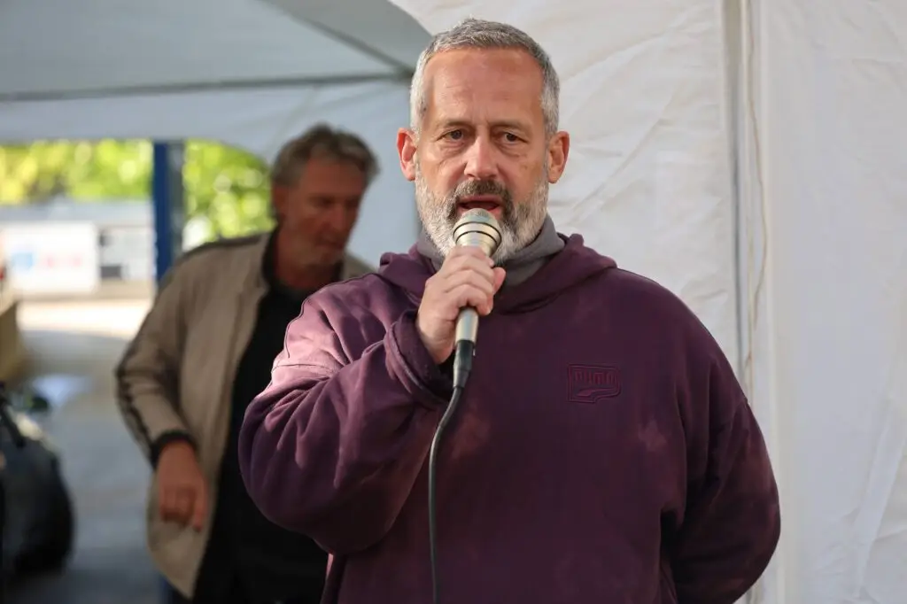 Man with gray beard and maroon hoodie speaks into a microphone under a white canopy.