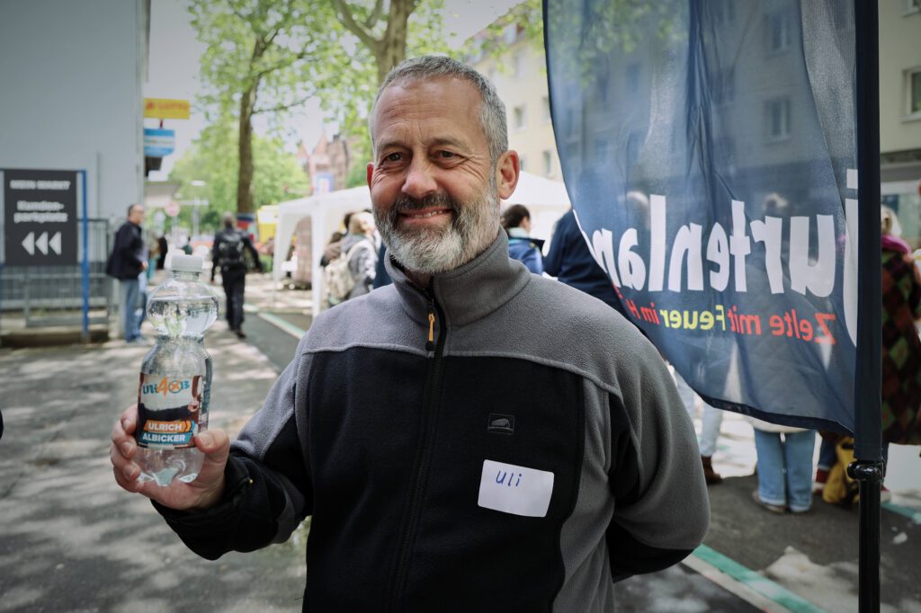 Smiling bearded man in a gray jacket at an outdoor event, holding a water bottle labeled Ulrich Albicker in the foreground.