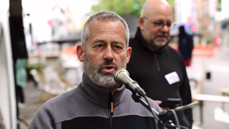 Ein Mann mit grauem Haar und Bart spricht in ein Mikrofon. Ein anderer Mann steht hinter ihm, unscharf.
