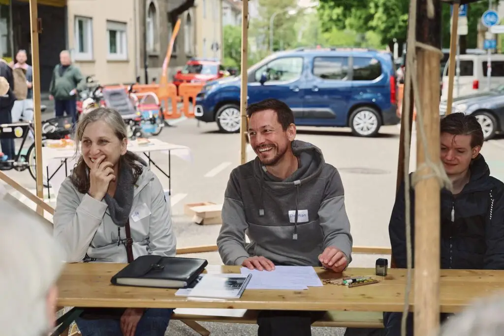 Drei Personen sitzen an einem Holztisch im Freien unter einem Vordach, lächeln und unterhalten sich. Auf dem Tisch liegen Papiere, ein Stift und ein Ordner. Hinter ihnen sind Autos und Baustellenabsperrungen auf der Straße zu sehen.