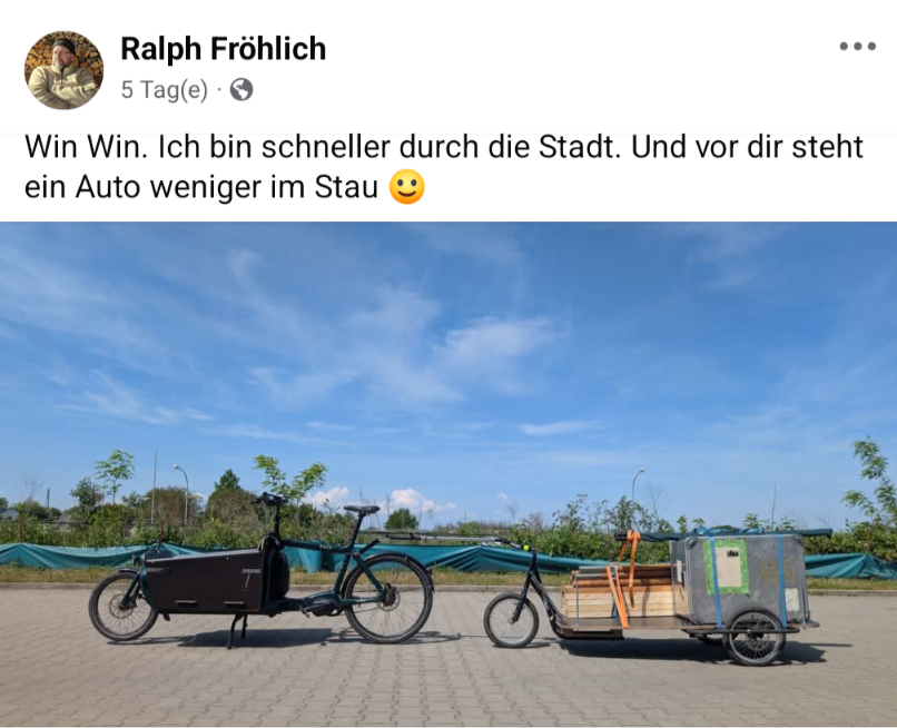 Ein schwarzes elektrisches Lastenfahrrad und ein mit Waren beladener Fahrradanhänger stehen auf einer gepflasterten Fläche unter blauem Himmel. Im Hintergrund sind Grünflächen und Bauzäune zu sehen.
