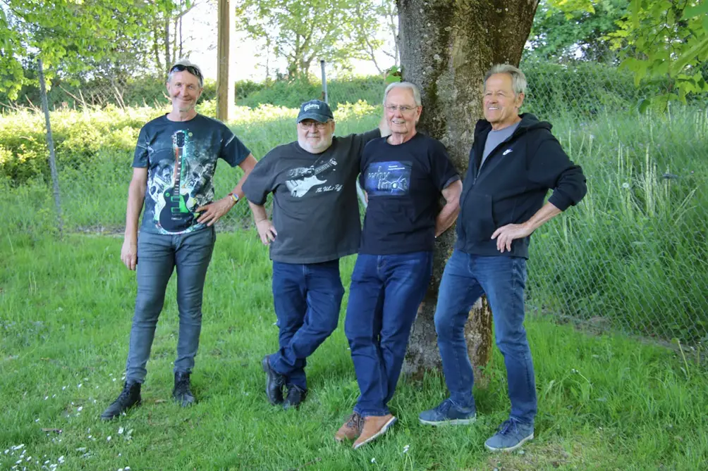 Vier Männer stehen lächelnd auf einer Wiese in der Nähe eines Baumes, mit Grünpflanzen und einem Drahtzaun im Hintergrund. Sie sind lässig in T-Shirts und Jeans gekleidet und genießen einen sonnigen Tag.
