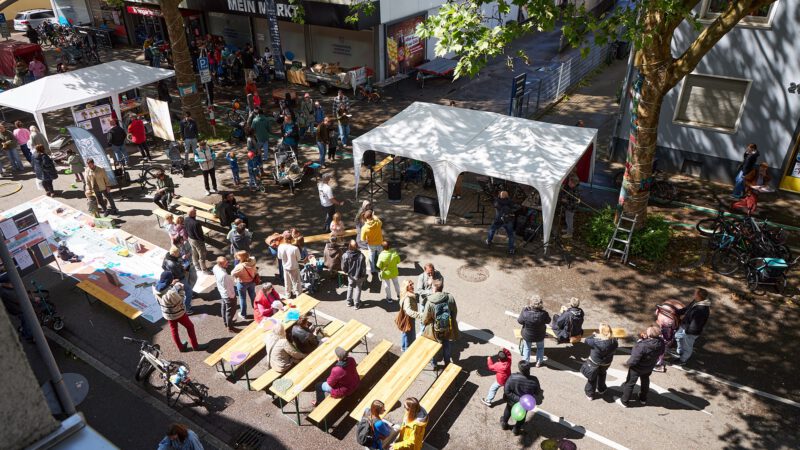 Busy street festival with white tents, tables, and crowds including families and kids enjoying activities.