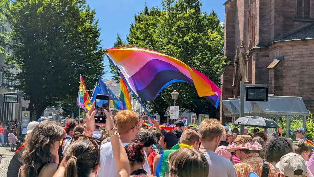 Eine Menschenmenge versammelt sich im Freien bei einer Pride-Parade und schwenkt verschiedene LGBTQ+-Fahnen, darunter eine große lesbische Pride-Flagge. Die Menschen feiern an einem sonnigen Tag in der Nähe eines historischen Gebäudes und von Bäumen.