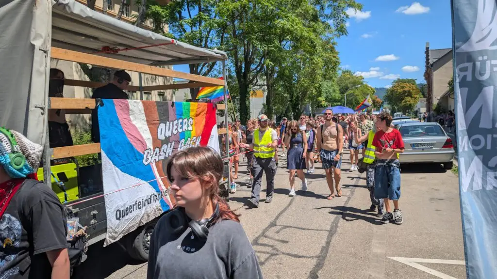 Eine farbenfrohe Pride-Parade mit Menschen, die eine sonnige Straße entlanglaufen. Ein mit einem Regenbogenbanner mit der Aufschrift "QUEER CASH LAB" geschmückter Lastwagen führt die Menge an. Einige tragen leuchtende Westen, andere haben Fahnen dabei, und viele wirken fröhlich und feierlich.