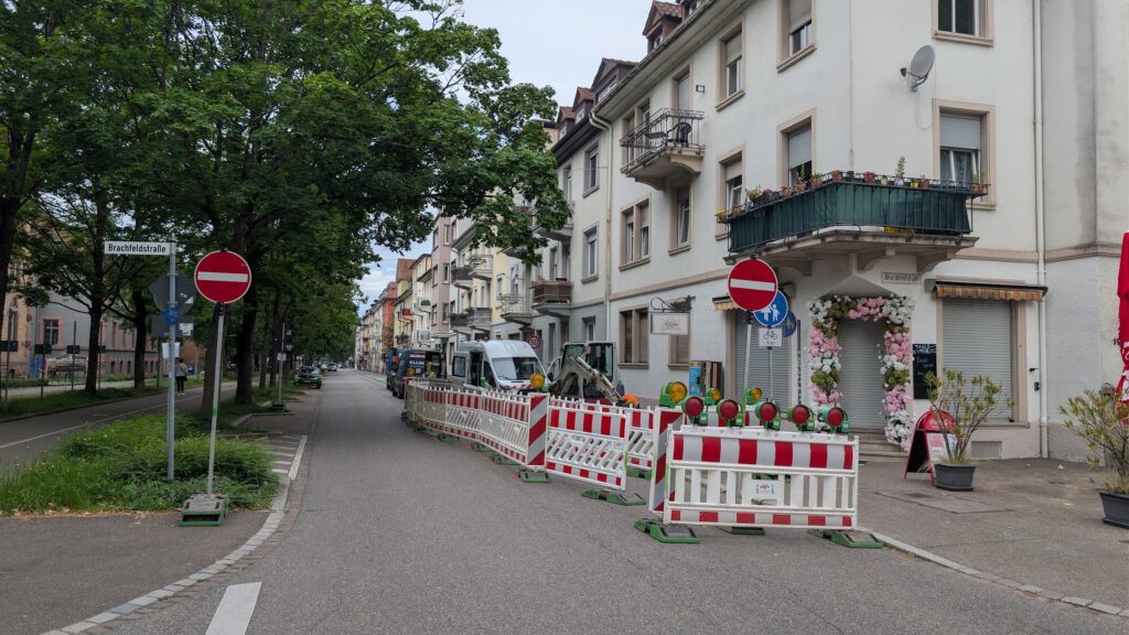 Street view with construction barriers along a residential block, trees on the left and a white building with a flower-filled balcony on the right.
