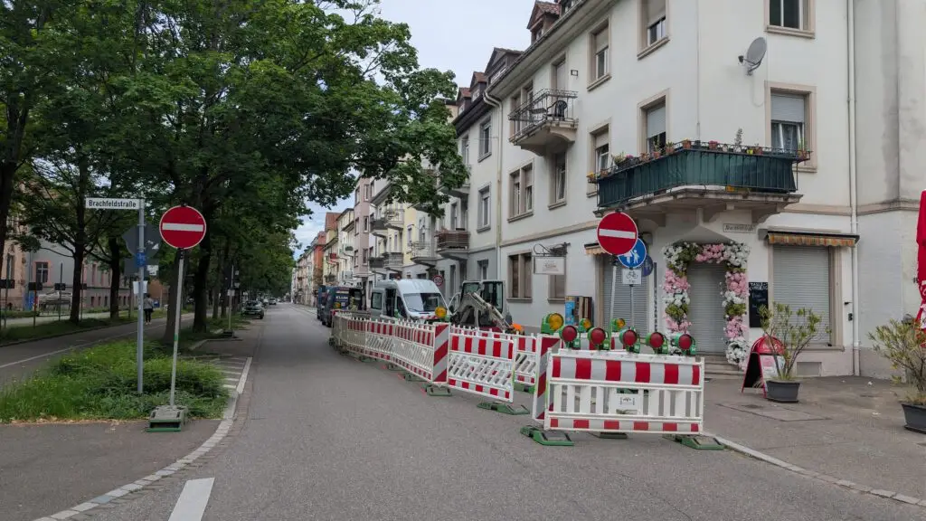 Street view with construction barriers along a residential block, trees on the left and a white building with a flower-filled balcony on the right.