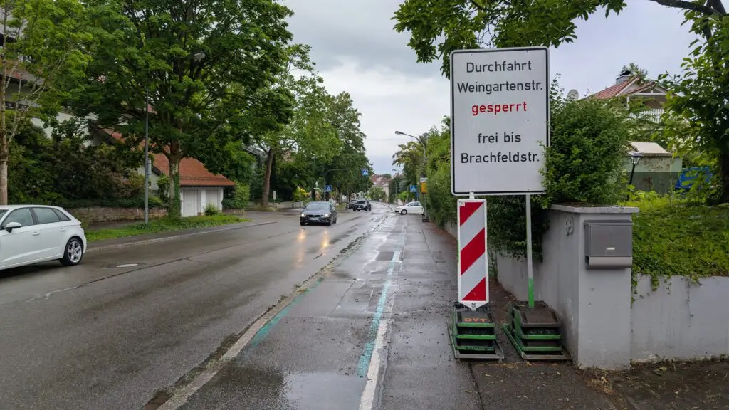 Wet street with a large road closure sign in German: 'Durchfahrt Weingartenstr. gesperrt frei bis Brachfeldstr.' indicating closed, with construction barriers nearby