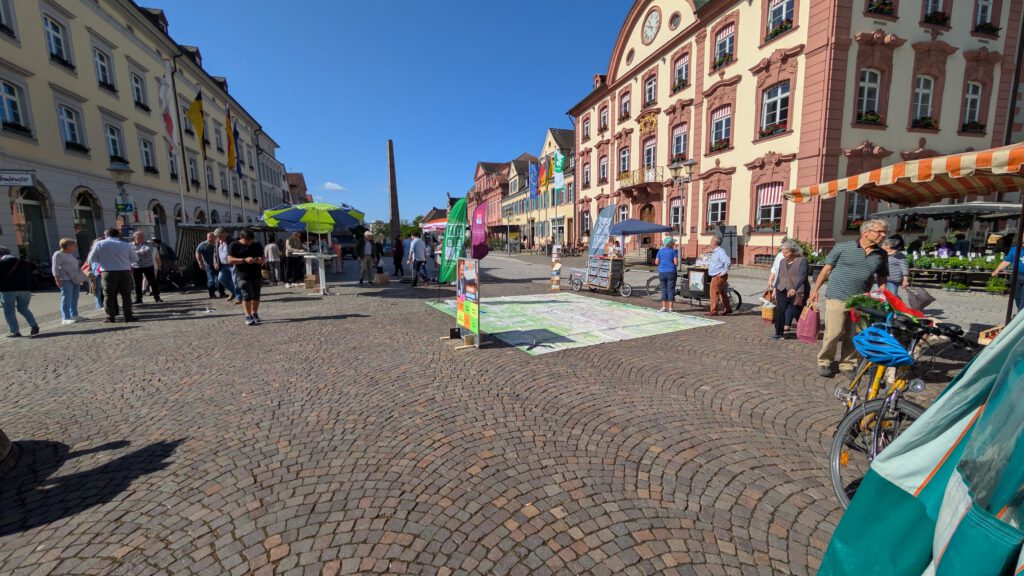 Die Menschen schlendern über einen Markt unter freiem Himmel auf einem mit Kopfsteinpflaster gepflasterten Platz, der von farbenfrohen historischen Gebäuden gesäumt ist, und stöbern an den Ständen. Auf dem Boden ist eine große Karte ausgebreitet, und in der Nähe stehen unter strahlend blauem Himmel Fahrräder.