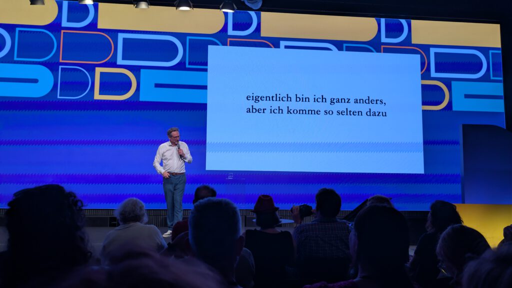 Ein Mann steht auf der Bühne vor einer Leinwand, auf der der deutsche Text „eigentlich bin ich ganz anders, aber ich komme so selten dazu“ zu sehen ist, und spricht zu den Zuschauern in einem hell beleuchteten, modernen Saal.