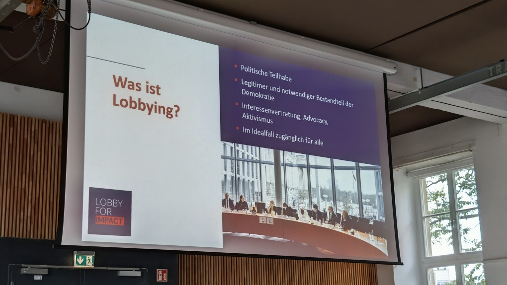 Eine Präsentationsfolie auf Deutsch mit dem Titel „Was ist Lobbying?“ listet Punkte zum Thema Lobbying auf und zeigt ein Foto von Menschen an einem Konferenztisch. Der Raum verfügt über Holzvertäfelung, ein Fenster und eine hohe Decke.