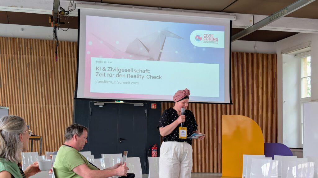 Eine Frau mit einem rosa Kopftuch spricht bei einer „Civic Coding“-Veranstaltung vor einer Projektionsfläche mit dem Titel „KI & Zivilgesellschaft – Zeit für den Reality-Check“ in ein Mikrofon. Mehrere Personen sitzen da und hören zu.
