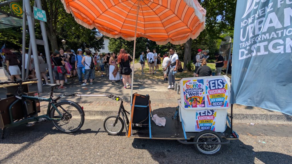 Ein Fahrradwagen mit einem großen, orange gestreiften Sonnenschirm verkauft bunte Eis am Stiel, die mit farbenfrohen Plakaten verziert sind. Bei einer Veranstaltung im Freien versammeln sich die Menschen in der Nähe unter Bäumen und einem blauen Banner.