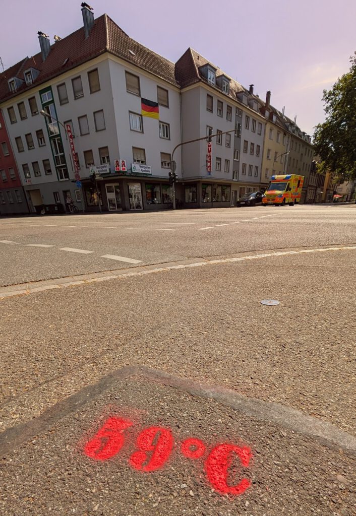 Auf dem Asphalt steht in roter Sprühfarbe „59 °C“. Im Hintergrund sind unter strahlend blauem Himmel eine Straßenecke mit einem Gebäude, Geschäften, einer deutschen Flagge und einem am Straßenrand geparkten Krankenwagen zu sehen.