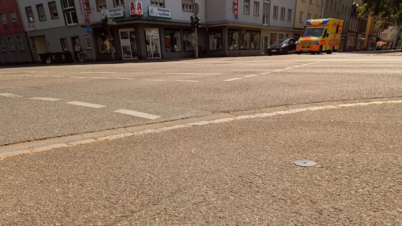 Auf dem Asphalt steht in roter Sprühfarbe „59 °C“. Im Hintergrund sind unter strahlend blauem Himmel eine Straßenecke mit einem Gebäude, Geschäften, einer deutschen Flagge und einem am Straßenrand geparkten Krankenwagen zu sehen.