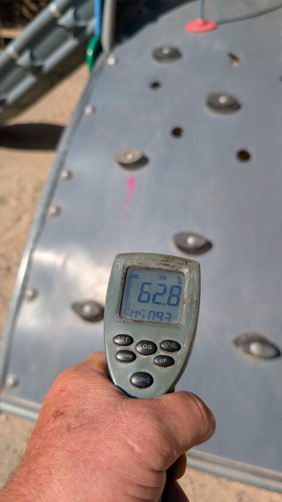 Eine Hand hält ein Infrarot-Thermometer, das 62,8 °C anzeigt, vor einer grauen Metallkletteranlage mit Haltegriffen auf einem Spielplatz. Im Hintergrund sind Sand und Spielgeräte zu sehen.