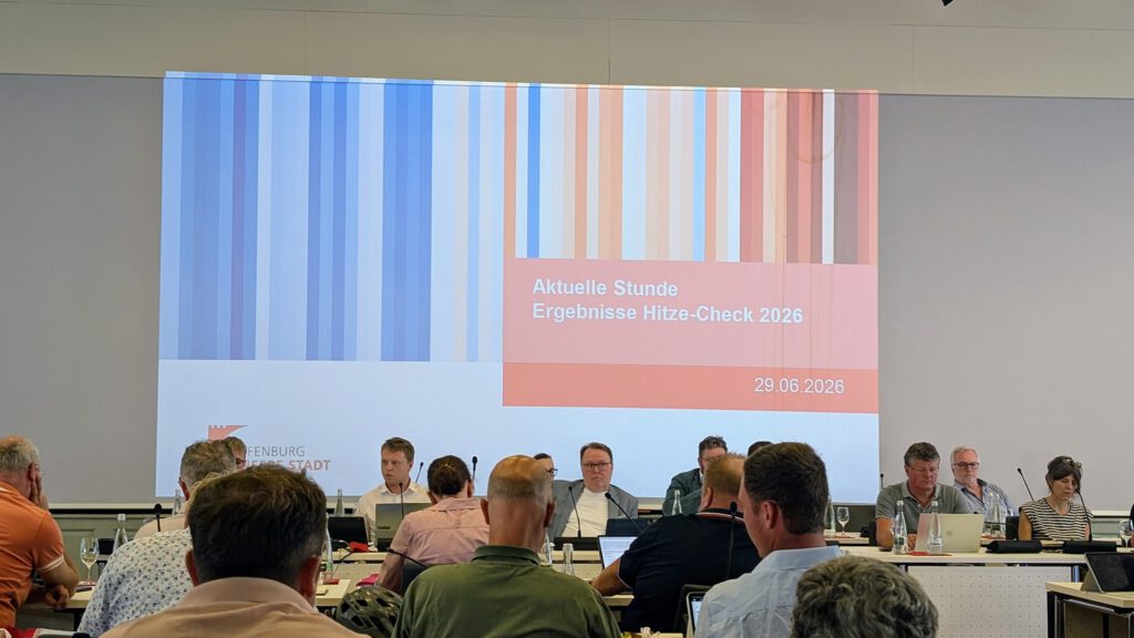 Ein Konferenzraum, in dem die Teilnehmer einem Podium mit Rednern gegenüber sitzen. Auf der Leinwand ist eine Folie mit der Aufschrift „Aktuelle Stunde – Ergebnisse Hitze-Check 2026, 29.06.2026“ zu sehen, die mit vertikalen Streifen in Blau, Rot und Weiß versehen ist.
