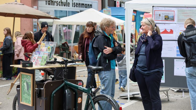 Outdoor street market with a green cargo bicycle selling magazines; people chat near tents and display boards in the background.