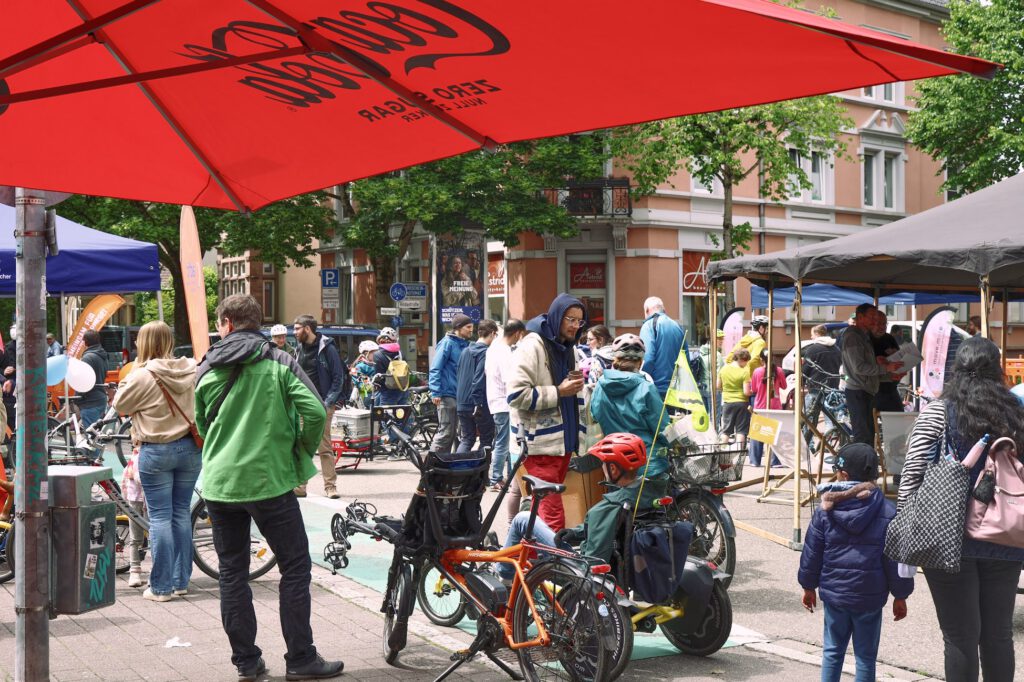 Unter großen Sonnenschirmen versammeln sich Menschen aller Altersgruppen, darunter auch Kinder mit Fahrrädern und Helmen, zu einer belebten Straßenveranstaltung. Stände und Transparente säumen die Straße, im Hintergrund stehen Bäume und Gebäude.