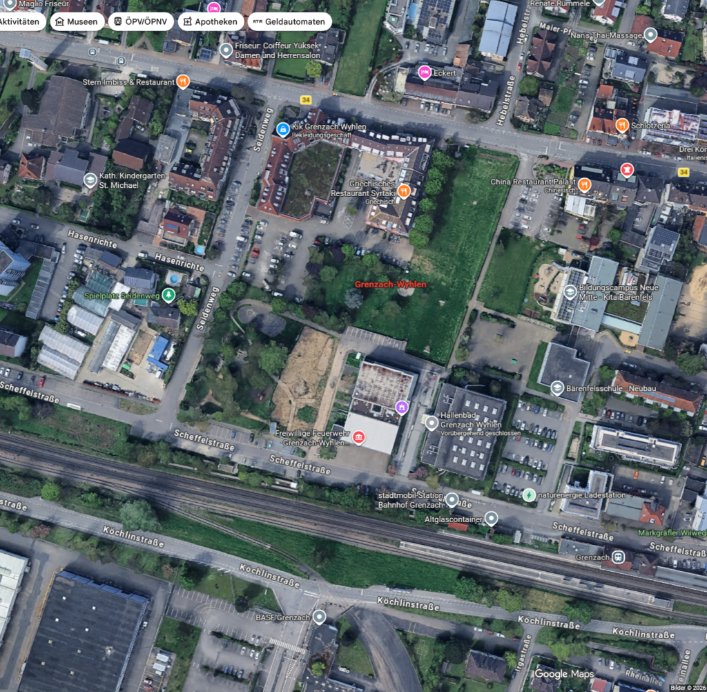 Luftaufnahme eines Stadtgebiets in Deutschland, auf der Straßen, Gebäude, Parkplätze, Grünflächen sowie beschriftete Orte wie Apotheken, Museen und Kindergärten zu sehen sind. Am unteren Bildrand verlaufen Bahngleise.