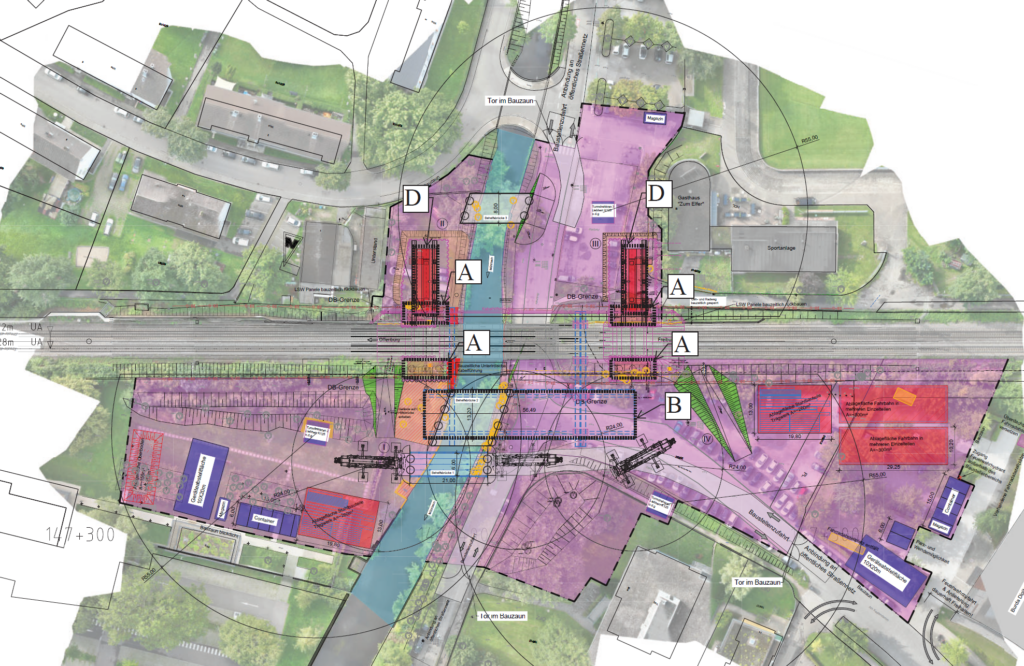 Ein detaillierter Baustellenplan, der über ein Luftbild gelegt ist, zeigt Gebäude, Straßen, Grünflächen sowie als Baustellenbereiche gekennzeichnete Stellen in Lila und Rot, wobei die Abschnitte mit A, B und D markiert sind und eine Eisenbahnlinie durch die Mitte verläuft.