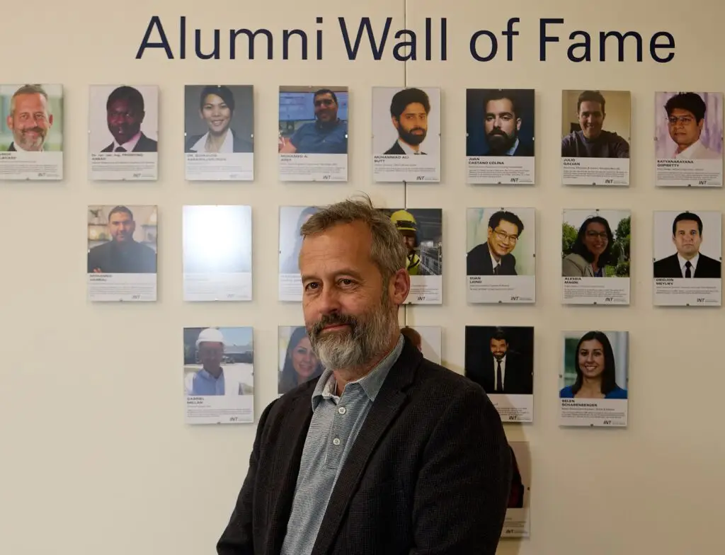 Ein Mann mit grauem Haar und Bart steht vor einer Fotowand mit der Aufschrift Alumni Wall of Fame. Hinter ihm sind mehrere Porträts von Alumni mit Namen und kurzen Beschreibungen zu sehen.